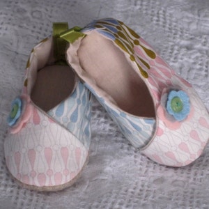 K&ouml;nnte beinhalten: Ein Paar Babyschuhe mit einem rosa, blauen und wei&szlig;en Blumenmuster. Die Schuhe haben einen Knopf vorne und ein gr&uuml;nes Band.