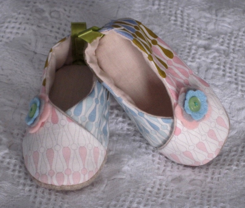 K&ouml;nnte beinhalten: Ein Paar Babyschuhe mit einem rosa, blauen und wei&szlig;en Blumenmuster. Die Schuhe haben einen Knopf vorne und ein gr&uuml;nes Band.