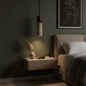 May include: A modern bedroom scene featuring a hanging pendant light with a dark wood top and a textured, cylindrical concrete base. A floating wooden nightstand holds a vase with dried pampas grass, books, and a diffuser. A framed line art print hangs above the bed.