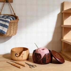 Peut inclure: Deux bols &agrave; fil en bois, un clair et un fonc&eacute;, avec un motif en spirale. Le bol fonc&eacute; contient de la laine rose et un crochet. Une paire de ciseaux et deux crochets sont devant les bols. Un panier avec une couverture &agrave; rayures bleues et blanches est en arri&egrave;re-plan.