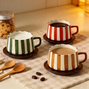 Op de afbeelding: Drie keramische mokken met strepen in groen, rood en oranje, elk op een houten onderzetter. Eén mok bevat melk, een andere koffie en de derde koffie. Een pot met koekjes en houten lepels zijn ook zichtbaar.