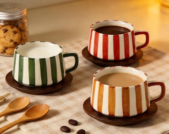 May include: Three ceramic mugs with striped patterns in green, red, and orange, each on a wooden coaster. One mug contains milk, another coffee, and the third coffee. A jar of cookies and wooden spoons are also visible.