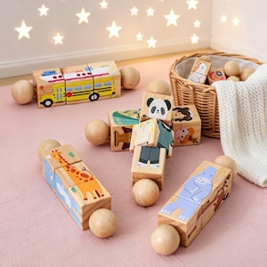 May include: Wooden toy blocks featuring animal and vehicle illustrations. A yellow school bus is assembled from blocks, along with blocks depicting a giraffe, hippo, and panda. The blocks are on a pink surface, with a basket and star-shaped lights in the background.