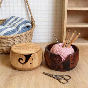 Peut inclure: Deux bols &agrave; fil en bois, un clair et un fonc&eacute;, avec une pelote de laine rose et des aiguilles &agrave; tricoter. Le bol clair porte l'inscription "Peace, Love, & Yarn Handcrafted for You". Une paire de ciseaux et un panier avec une couverture &agrave; rayures bleues et blanches sont &eacute;galement pr&eacute;sents.