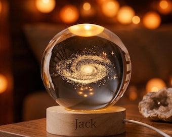 Peut inclure: Une boule de cristal transparente avec un motif de galaxie illuminé. Elle repose sur un socle en bois gravé du nom "Jack". Un câble blanc part de la base. L'arrière-plan est flou avec des lumières chaudes, créant une ambiance chaleureuse.