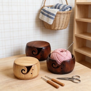 Peut inclure: Trois bols &agrave; fil en bois de diff&eacute;rentes nuances de marron et de bois naturel, chacun avec un guide &agrave; fil en forme de spirale. Un bol contient une pelote de laine rose avec un crochet. Deux crochets et des ciseaux sont &eacute;galement sur la table.