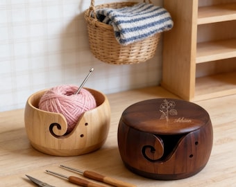 Peut inclure: Deux bols à fil en bois, des crochets et une paire de ciseaux sur une surface en bois. Un bol contient de la laine rose et un crochet. Un bol en bois foncé avec un motif floral et le nom "Adrian" est également présent. Un petit panier est suspendu au-dessus.