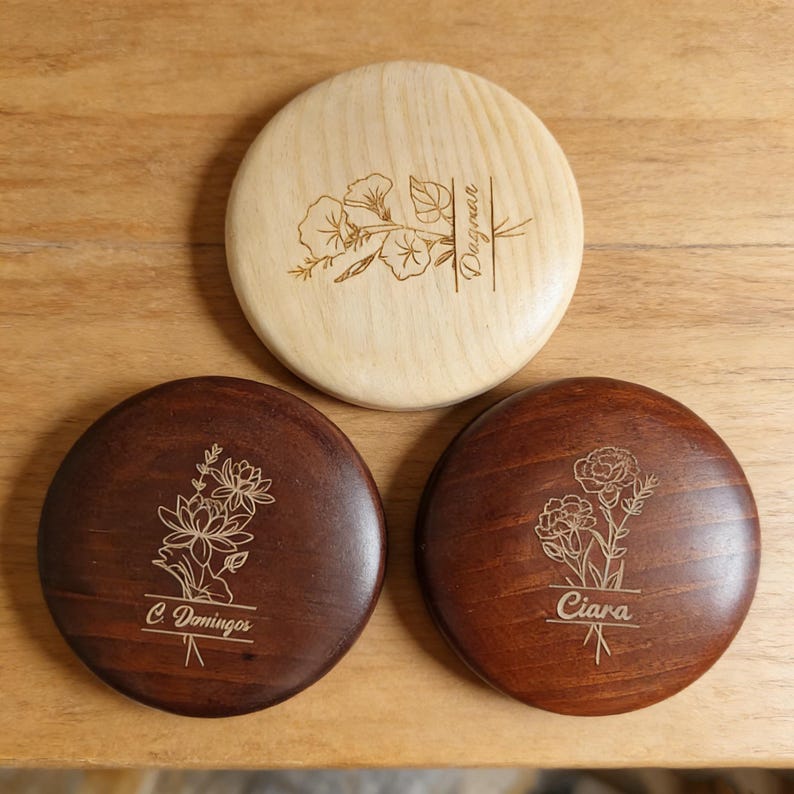 Peut inclure: Trois dessous de verre en bois ronds avec des motifs floraux grav&eacute;s. L'un est de couleur claire avec un motif floral et le nom "Dagnan". Les deux autres sont brun fonc&eacute;, l'un avec "C Domingos" et l'autre avec "Ciara".