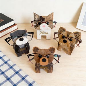 Puede incluir: Cuatro soportes de madera para gafas con forma de animales: un oso, un gato, un perro y un oso. Cada soporte tiene un par de gafas. El soporte para gatos tiene el nombre "Nora". El soporte para osos tiene el nombre "Adrian". El soporte para osos tiene el nombre "Lucas".