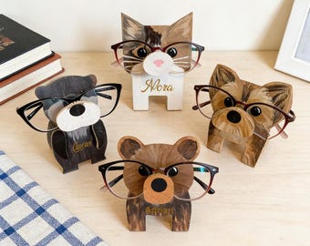 Peut inclure: Quatre supports à lunettes en bois en forme d'animaux : un ours, un chat, un chien et un ours. Chaque support porte une paire de lunettes. Le support pour chat porte le nom "Nora". Le support pour ours porte le nom "Adrian". Le support pour ours porte le nom "Lucas".