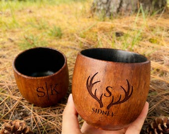Peut inclure: Deux gobelets en bois avec un intérieur sombre. L'un porte les initiales "SK", l'autre un motif de bois de cerf avec le nom "SIDNEY" et la lettre "S". Les gobelets sont bruns et posés sur des aiguilles de pin.