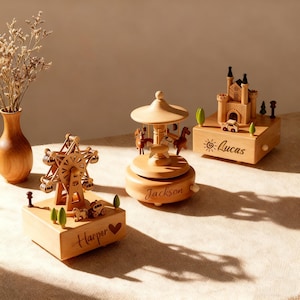 May include: Three wooden music boxes with intricate designs. One features a Ferris wheel, another a carousel with horses, and the third a castle. Each box has a name engraved on it: "Harper", "Jackson", and "Lucas".