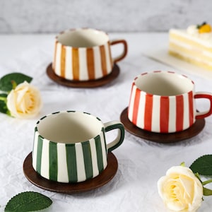 Könnte beinhalten: Drei Keramikbecher mit vertikalen Streifen in Orange, Rot und Grün, jeweils auf einem kleinen Holzuntersetzer. Die Becher haben farblich passende Henkel. Der Hintergrund ist weiß, mit einem Stück Kuchen und Blumen.