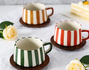May include: Three ceramic mugs with vertical stripes in orange, red, and green, each on a small wooden coaster. The mugs have matching colored handles and are set against a white background with a slice of cake and flowers.
