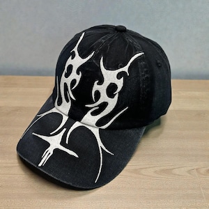 Może przedstawiać: Czarna czapka bejsbolówka z białym haftem w kształcie plemiennych płomieni z przodu i na daszku. Czapka ma zakrzywiony daszek i guzik na górze. Wzór haftu jest symetryczny i pokrywa przód czapki.