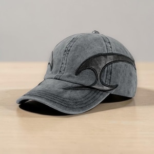 Może przedstawiać: Szara czapka bejsbolowa z zakrzywionym czarnym skórzanym wzorem z boku. Czapka ma zakrzywiony daszek i metalowy guzik na górze. Tkanina ma zużyty, wyblakły wygląd.