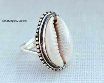 May include: A silver ring featuring a natural cowrie shell centerpiece. The shell is a creamy white color, set within a beaded silver border. The ring band splits into two at the base of the shell. The text "BohoMagic925Jewel" is visible.