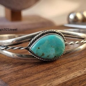May include: A silver cuff bracelet featuring a central pear-shaped turquoise stone with a rope-style border. The band consists of two smooth silver wires and one twisted silver wire. The text BohoMagic925Jewel is visible in the background.