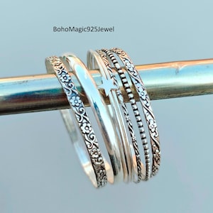 Puede incluir: Una colección de pulseras de plata con varios diseños, incluyendo patrones florales y bandas texturizadas. Las pulseras se muestran en una barra de plata sobre un fondo azul claro. Joyería adecuada para el uso diario.