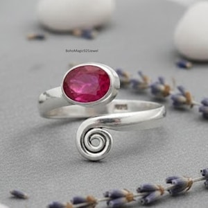 May include: A silver ring featuring a large, oval, ruby-coloured gemstone. The ring has a distinctive design with a spiral detail. The ring is presented on a grey surface with lavender sprigs and white stones.