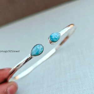 May include: A silver-toned cuff bracelet with two teardrop-shaped, light blue gemstones. The open design features a hammered texture on the band. The text "Bohomagic925Jewel" is visible.
