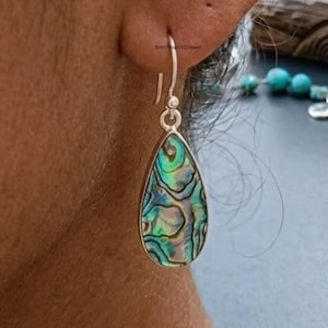 May include: Abalone shell teardrop earrings with a silver hook. The iridescent shell displays shades of green, blue, and purple. The earrings are a unique piece of jewelry. The earrings are shown on a person's ear.