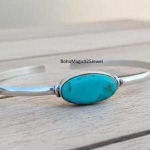 Könnte beinhalten: Ein silbernes Armreif-Armband mit einem leuchtend türkisfarbenen, ovalen Stein in der Mitte. Das Armband hat ein schlichtes, elegantes Design, perfekt für den täglichen Gebrauch. Der Türkisstein weist natürliche Farbvariationen auf.