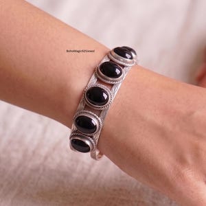 Könnte beinhalten: Ein silberner Armreif mit fünf ovalen schwarzen Steinen, die in gedrehten Metallfassungen sitzen. Das Band ist an den Seiten mit einem Blattmuster graviert. Der Text BohoMagic925Jewel ist sichtbar. Das Design ist offen gestaltet.