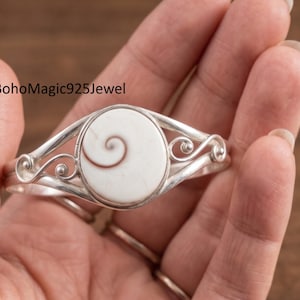 May include: A silver cuff bracelet featuring a central oval-shaped white shiva eye shell with a brown spiral pattern. The metal band displays decorative swirling wire details on both sides of the stone setting. The text BohoMagic925Jewel is visible.