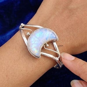 Puede incluir: Una pulsera de plata con una piedra lunar iridiscente en forma de media luna engastada en el centro. La banda de metal tiene un diseño dividido con pequeños acentos esféricos en los puntos donde el marco se conecta con el engaste central.