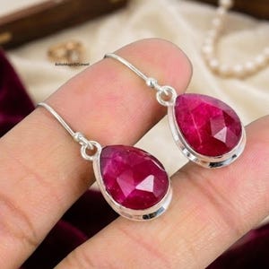 May include: Silver-tone drop earrings with faceted, teardrop-shaped, ruby-red gemstones. The earrings have a simple, elegant design, with the gemstones set in a silver bezel. The earrings are shown against a blurred background.