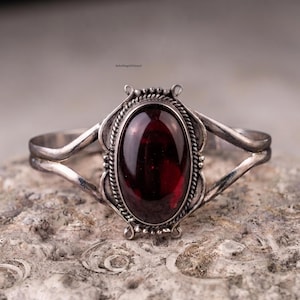 May include: A silver cuff bracelet featuring a large, oval, deep red gemstone at the centre. The stone is surrounded by a twisted rope-style metal border and decorative silver accents. The band splits into two thin, polished silver arms on each side.