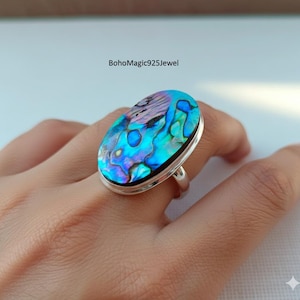 Puede incluir: Anillo de plata ovalado con una concha de abulón iridiscente en tonos azules, verdes y morados. El anillo tiene una banda de plata pulida y un engaste biselado. El texto "BohoMagic925Jewel" es visible.
