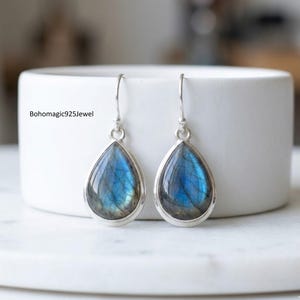 May include: A pair of teardrop-shaped earrings with iridescent blue and silver tones. The earrings have a silver frame and a hook closure. The earrings are displayed on a white surface. The text "Bohomagic925Jewel" is visible.