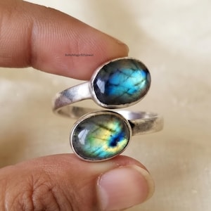 May include: A bypass ring featuring two oval-shaped labradorite gemstones set in a silver band. The top stone displays iridescent blue flashes, while the bottom stone shows a mix of yellow and green tones. The text BohoMagic925jewel is visible.