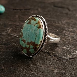 May include: A silver ring featuring a large, oval-shaped turquoise stone with brown matrix patterns. The stone is set in a twisted rope-style silver border. The band is split into two sections where it meets the setting. Text reads BohoMagic925Jewel.