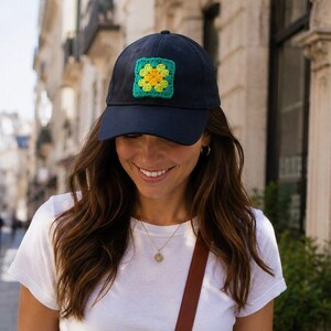 May include: A navy blue baseball cap featuring a square, crocheted patch on the front. The patch consists of a bright yellow centre surrounded by light green petals, all framed by a dark teal border, creating a floral-inspired square design.