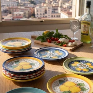 Può includere: Una collezione di piatti in ceramica con motivi di limoni gialli dipinti a mano, foglie verdi e bordi decorativi a volute in vari colori, tra cui blu, giallo e verde, disposti su un tavolo di legno con bicchieri.