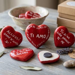 Op de afbeelding: Meerdere rode keramische harten met witte handgeschreven tekst in verschillende talen, waaronder zinnen zoals TI AMO, ACCUSSI SARRA' PE' SEMPRE SI en TU SI' NA COSA GRANDE PE' ME. Eén hart heeft een zwarte ronde magneet op de achterkant.