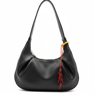 Peut inclure: Sac à bandoulière en cuir noir de forme incurvée avec une seule anse. Le sac est doté d'une fermeture éclair dorée et d'un pompon décoratif rouge et vert avec un anneau jaune. Le sac est posé sur un fond blanc.