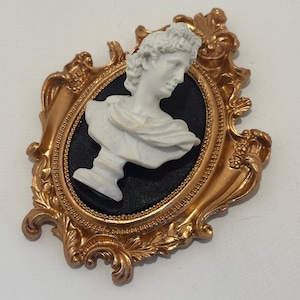 Könnte beinhalten: Eine goldfarbene, verzierte Brosche mit einer weißen Büste einer klassischen Figur vor schwarzem Hintergrund. Der Rahmen hat aufwendige Details und eine Vintage-Ästhetik. Die Brosche ist wahrscheinlich aus Metall und ein dekoratives Accessoire.
