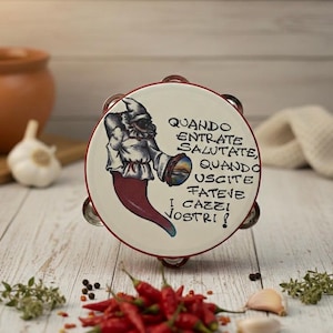 Può includere: Un tamburello in ceramica con bordo rosso e un disegno dipinto con un peperoncino rosso e una figura con camicia bianca. Il testo sul tamburello recita "QUANDO ENTRATE SALUTATE, QUANDO USCITE FATEVE I CAZZI VOSTRI!" Peperoncini rossi e aglio in primo piano.