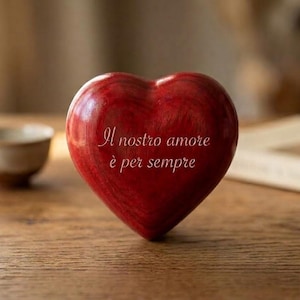 Op de afbeelding: Een glad, gepolijst rood hartvormig object met een zichtbare houtnerfstructuur. In het midden van het hart staat de gegraveerde witte tekst "Il nostro amore è per sempre", wat "Onze liefde is voor altijd" betekent.