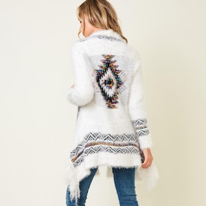 Puede incluir: Cárdigan blanco abierto con un diseño geométrico colorido en la espalda y el dobladillo. El suéter de manga larga tiene una textura difusa y se combina con jeans azules y botas altas marrones.