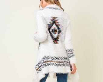 May include: White open-front cardigan with a colorful geometric design on the back and hem. The long-sleeved sweater has a fuzzy texture and is paired with blue jeans and brown knee-high boots.
