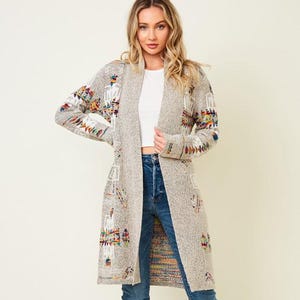 Könnte beinhalten: Lange, offen geschnittene Strickjacke in hellgrauem Strick mit bunten geometrischen Mustern auf den Ärmeln und der Vorderseite. Die Jacke wird mit blauen Jeans und braunen kniehohen Stiefeln getragen.