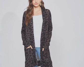 May include: A long, open-front cardigan in a dark gray leopard print. The cardigan has two front pockets and is worn over a white top and blue jeans. The model is wearing brown ankle boots.