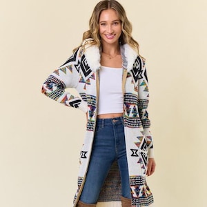 May include: A long, cream-colored cardigan with a geometric pattern in black, red, blue, and yellow. The cardigan has a faux fur collar and is paired with blue jeans and brown knee-high boots. The cardigan is a long length.