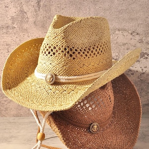 May include: Two straw cowboy hats stacked together, one light beige and one medium brown. Each hat features a woven texture, a white rope hatband, and a circular metal emblem displaying an anchor. A white chin cord with wooden beads hangs down.