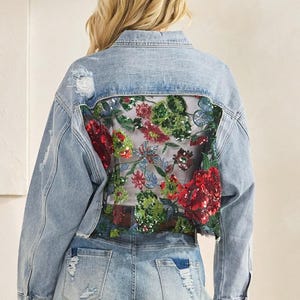 May include: Pale blue denim jacket with distressed detailing and a floral sequin design on the back. The jacket is paired with matching denim shorts. The shorts have a frayed hem and distressed detailing.